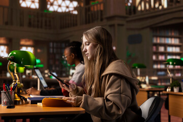 Young girl reading a literature book to collect citations for her bachelor paper, ensuring a plagiarism free diploma paperwork at the university library. Student takes notes on her smartphone.