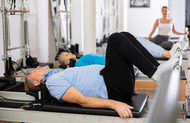Mature male pensioner in sportswear performing exercise lying down on reformer bed during back training in Pilates center