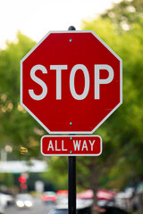 Red metal Stop All Way street sign on the side of a road, vertical, blurred green street background
