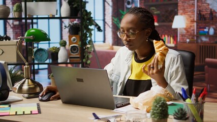 Busy freelancer in home office eating snack on the go while typing on laptop keyboard. Woman enjoying croissant while using electronic device to solve job tasks online, camera A