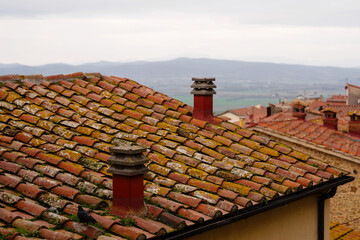 Close-up of weathered tile rooftop with chimneys and antenna. The distant rolling hills under cloudy sky offer backdrop to this rustic urban setting