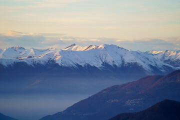 Misty valley lies beneath snow-capped mountain peaks at dusk near Piani di Bobbio in Italy, with soft light creating serene and tranquil alpine landscape