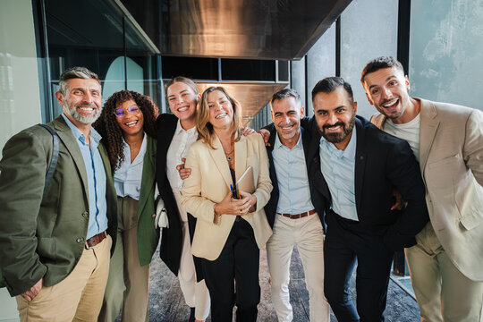 Diverse group of smiling business professionals standing together in office corridor, dressed in formal attire, celebrating teamwork, success, corporate unity and collaboration while posing happily
