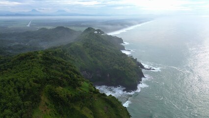 Aerial drone view of coastline with hills, cliffs, trees, beach sand and waves from the sea and ocean at Karang Bolong Beach, Kebumen, Central Java, Indonesia