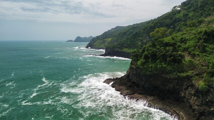 Aerial drone view of coastline with hills, cliffs, trees, beach sand and waves from the sea and ocean at Karang Bolong Beach, Kebumen, Central Java, Indonesia