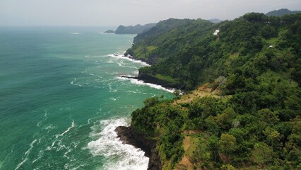 Fototapeta premium Aerial drone view of coastline with hills and trees, as well as view of coral cliffs and sea with waves from the ocean in Sagara view also known as Karang Bolong Beach Kebumen Central Java Indonesia
