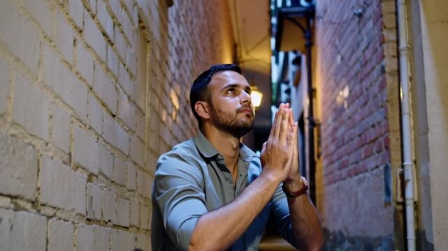 Man sitting in a narrow alley, hands clasped in prayer, seeking guidance and resilience in challenging times.