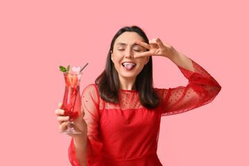 Beautiful young woman holding glass of tasty cocktail with strawberry and showing victory gesture on pink background