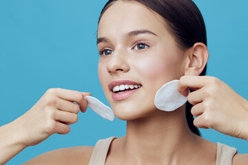 Young woman with smooth skin applying cotton pads to her face, showcasing a glowing complexion, joy, and beauty against a bright blue background