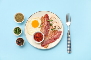 Plate with tasty fried egg, bacon, mushrooms and spices in bowls on blue background. Top view