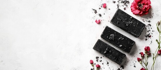 Black soap bars on white background surrounded by pink flowers and herbs with Copy Space for text placement.