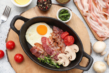 Pan with tasty fried egg, bacon, mushrooms, tomatoes and spices in bowls on wooden stand on white grunge background. Top view