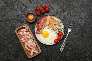 Raw egg, bacon, plate with fried egg, bacon, toasts, mushrooms and tomatoes on black background. Top view
