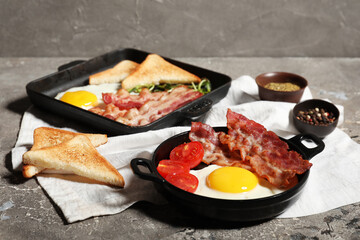 Two portions of fried eggs, bacon, toasts and bowls of spices on table against grey background
