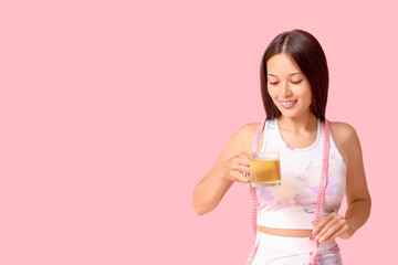 Beautiful young happy Asian woman in sportswear with measuring tape and cup of tea for weight loss on pink background