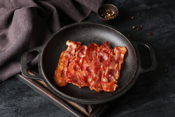 Frying pan with fried bacon rashers and bowl of peppercorn on black background