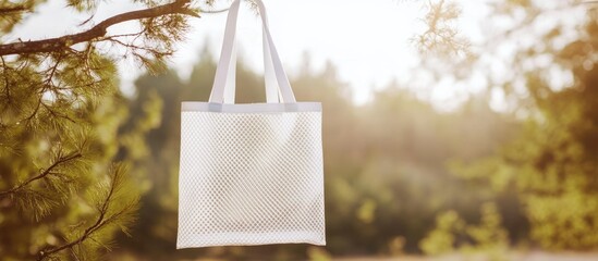 White mesh tote bag hanging from a tree branch in a natural setting with soft sunlight and blurred trees creating space for your text design
