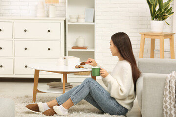 Fototapeta premium Beautiful young Asian woman with cup of hot tea and sweet cookies sitting on carpet in living room