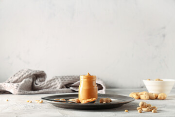 Jar of tasty peanut butter with spoon and nuts on light background