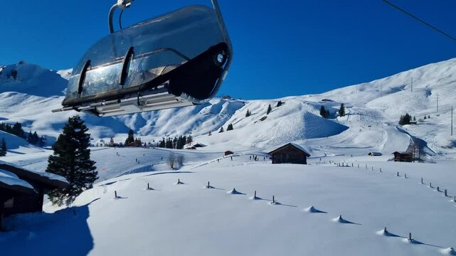 Panoramic view of snow-covered alpine mountains with chalets and people skiing on the distant slopes, filmed from a moving chairlift on a sunny winter day in Adelboden Lenk Switzerland