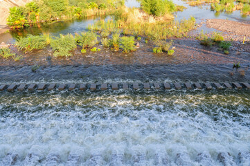 Flowing water over a stepped dam surrounded by lush vegetation and rocky terrain, depicting water management and ecological balance in the Rio Salado en Tempe, Arizona