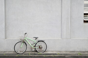 A pastel green bicycle leans gently against a gray concrete wall, resting atop a galvanized steel drainage grate. The muted tones and simple composition highlight a quiet urban moment.