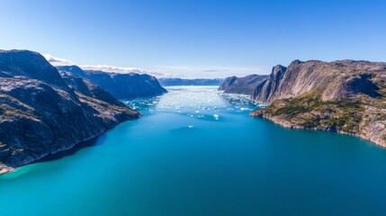 Obraz premium Aerial view of a fjord in Greenland, showcasing icebergs floating in turquoise waters between towering mountains under a clear sky, ideal for travel brochures