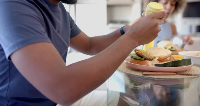 Adding mustard to burger, man enjoying meal with Diverse friends at table