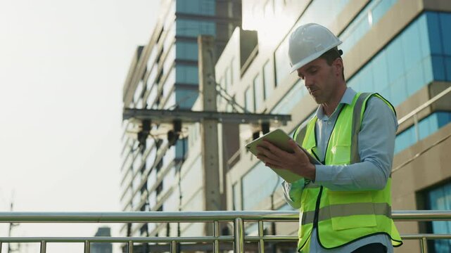 Man construction engineer examines project documentation on tablet against factory building. Male worker wearing hardhat and safety vest ensures precise execution of work from footbridge