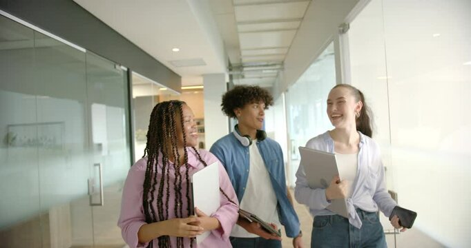 Walking in hallway, three students carrying laptops and tablets, smiling and talking