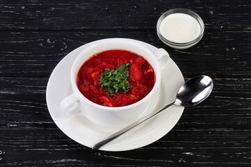 Traditional Ukrainian and Russian borscht in a white ceramic plate with herbs, sour cream and garlic on a black wooden background