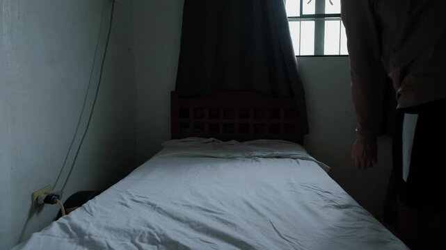 Young man fixing bed sheets on twin size bed in sunlit bedroom -static wide shot