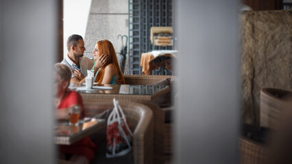 couple sitting at a table in a cafe or restaurant. The man and woman look at each other with smiles. The man is wearing a light shirt, and the woman has long red hair.