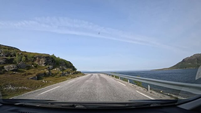 Driving through the tundra on E69 in northern Norway. Rocky hills with sparse trees and grass rise on the left, while the sea stretches along the right, showcasing raw and rugged nature.