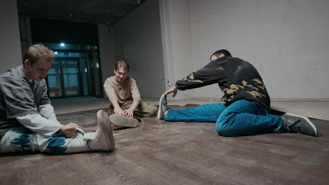 Ground level shot of three Caucasian male zoomers wearing street style clothes stretching legs on dance floor, talking while warming up before training indoors