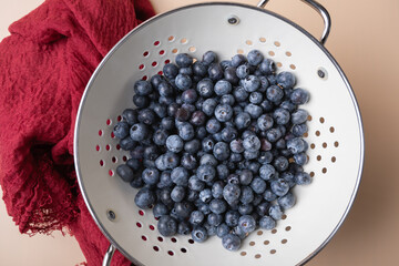 Fresh blueberries in white metal strainer next to red cloth on beige background, combining naturalness and elegance. Healthy eating concept