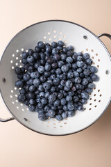 Fresh blueberries in white colander with metal handles on beige background, highlighting nutrition and freshness. Concept of natural and healthy food