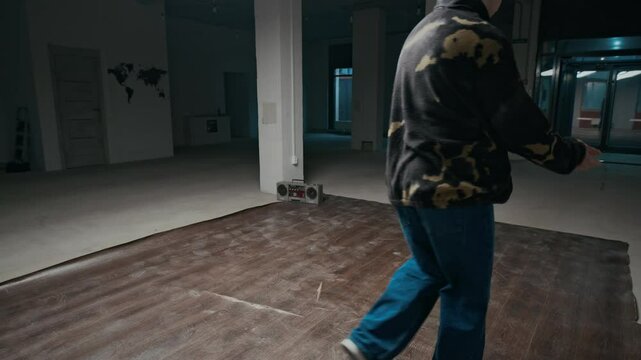 Stab shot of gen Z Caucasian b-boy in streetwear approaching camera, start recording his breakdance performance with top rock and down rock elements, training in empty dance hall indoors