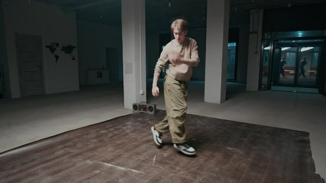 Stab shot of talented male Caucasian zoomer wearing hip hop clothes approaching camera and recording himself breaking, performing top rock and down rock moves while practicing in empty indoor hall