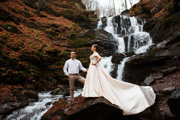 couple standing against the backdrop of a waterfall. Nature surrounds them: stones covered with moss and falling water.