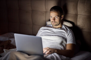 A young man is using laptop in bed late at night in dark bedroom.