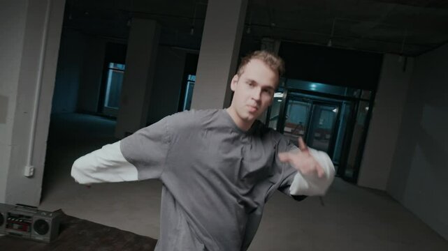Handheld shot of inspired Caucasian male zoomer in street style clothes performing breakdance moves of excellent footwork, freestyle, improvising and enjoying hip hop indoors