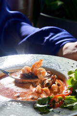 A woman's hand reaches for a steaming bowl of Tom Yum soup, filled with succulent shrimp, fragrant herbs, and a fiery red broth.