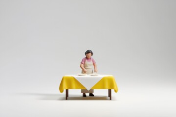 A miniature figurine of a woman setting a yellow tablecloth table with plates.