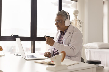 Virtual checkup. Busy older age doctor in earphones sit at desk focused on notebook screen communicate with patient by videocall discuss disease symptoms make diagnosis prescribe treatment by internet
