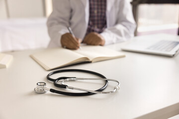 After seeing patients. Close up male physician in charge work with paper documents in hospital office fill in information in register journal on duty shift. Focus on stethoscope on doctors work desk
