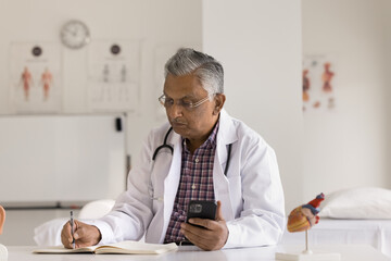 Smartphone and medicare. Busy mature male doctor medic worker physician use modern mobile phone at desk in hospital office make schedule of appointments take notes on paper from message text on screen
