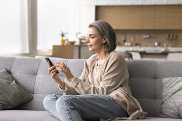 Obraz premium Exploring digital world. Happy mature female engaged in virtual communication on cell at home type email text send message to friend at chat book services choose buy goods online using smartphone app