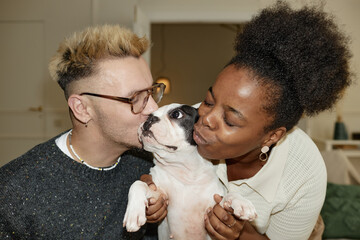Portrait of excited Black woman and her boyfriend hugging and kissing little French bulldog sharing love and affection for little pet captured at home, camera flash