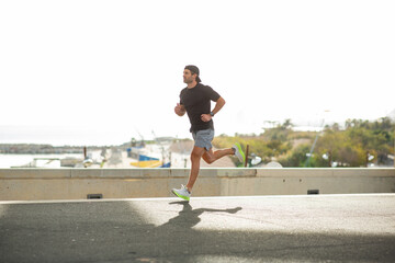 Man in black t-shirt, cap, gray shorts and white sneakers running outdoors by the sea during a...